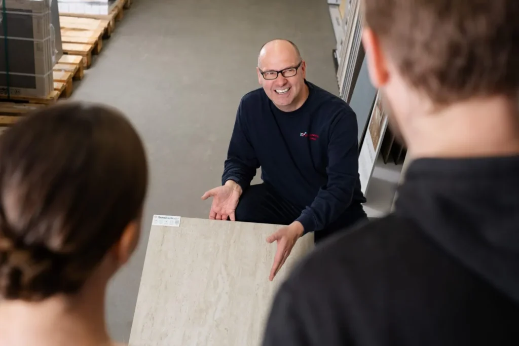 Mitarbeiter zeigt Fliesenplatte während Beratungsgespräch mit zwei Kunden – Business-Foto unserer Partnerfotografen