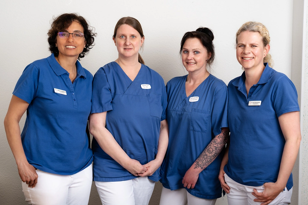 Teamfoto einer Arztpraxis oder Pflegeeinrichtung – vier Mitarbeiterinnen in blauer Arbeitskleidung vor weißer Wand